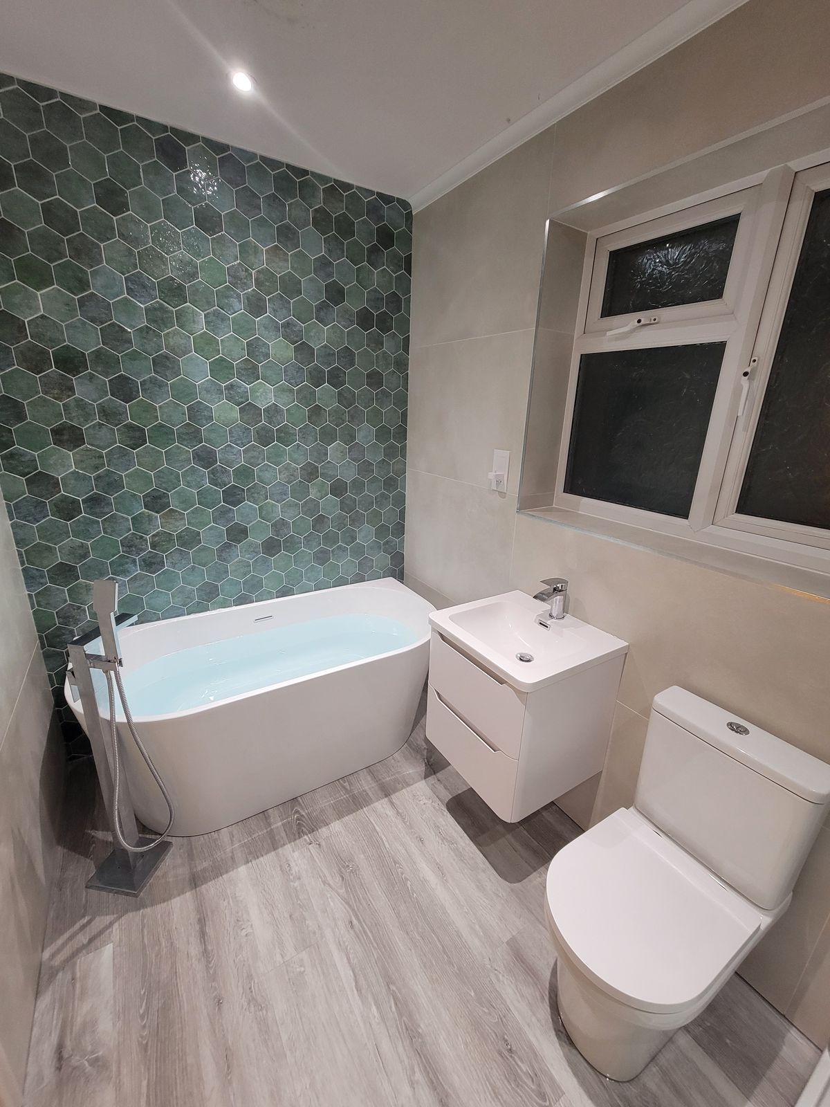 A freestanding bath against a wall of green hexagon tiles, with a wall-hung basin unit