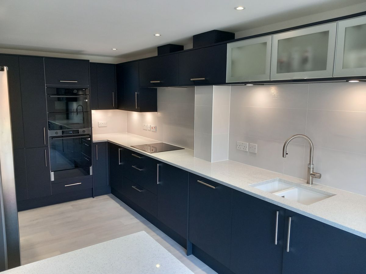 A dark navy handleless kitchen with white worktops, built-in ovens and glass wall units
